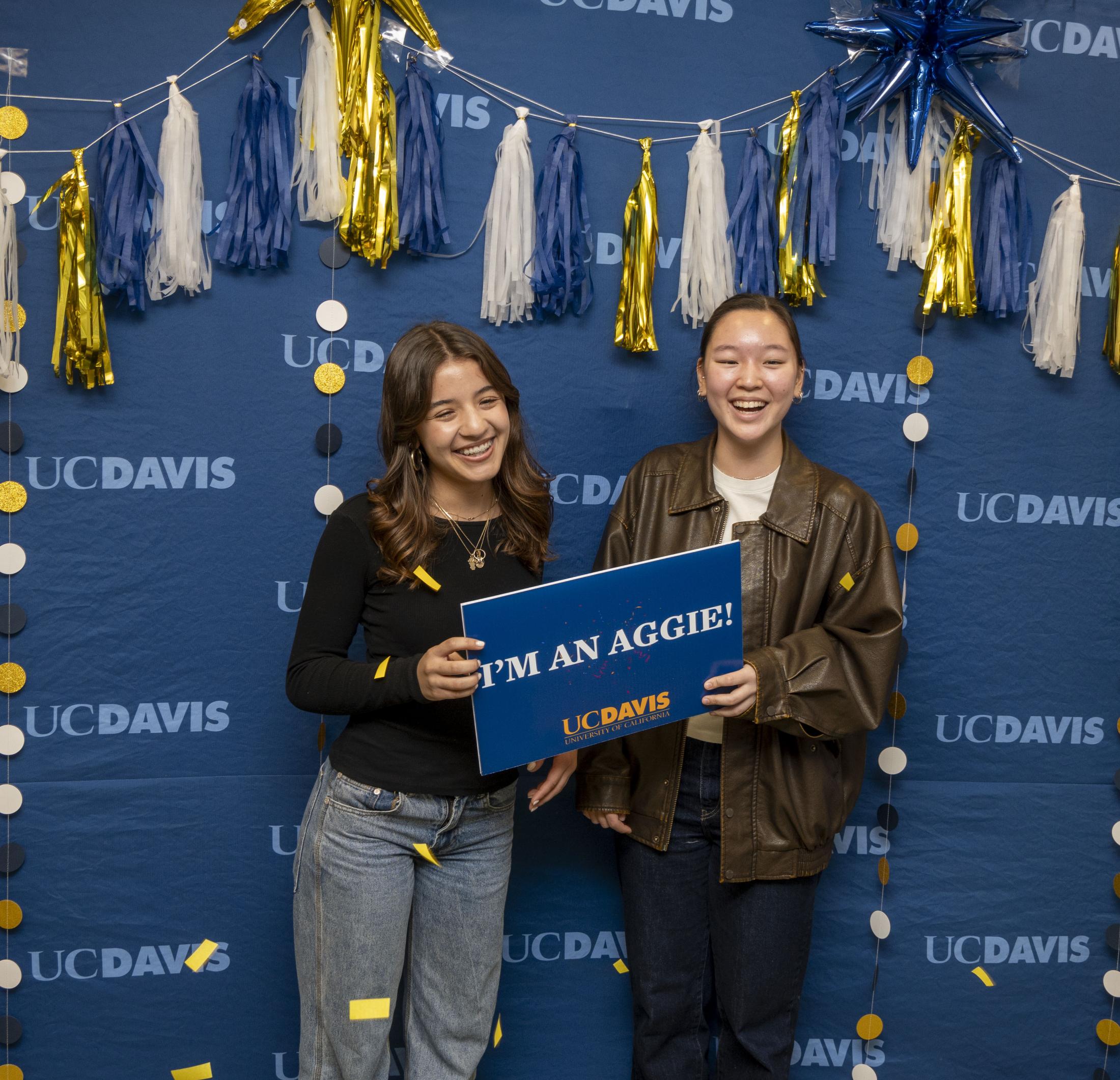 Two UC Davis admits posing for a photo in front of a celebratory backdrop.