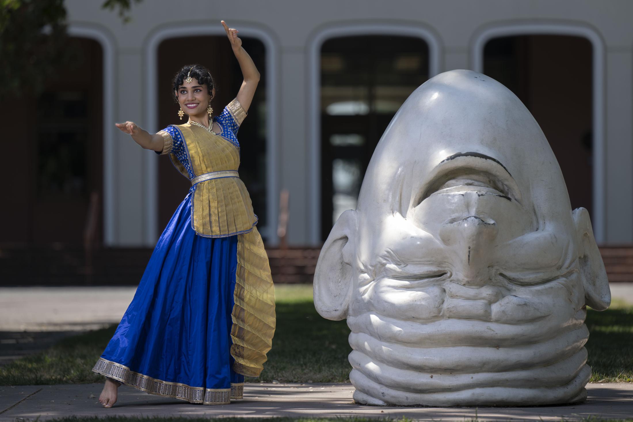 A young woman in a vibrant blue and gold traditional Indian dance costume poses barefoot in front of Egghead sculpture, Eye on Mrak, on the UC Davis campus.