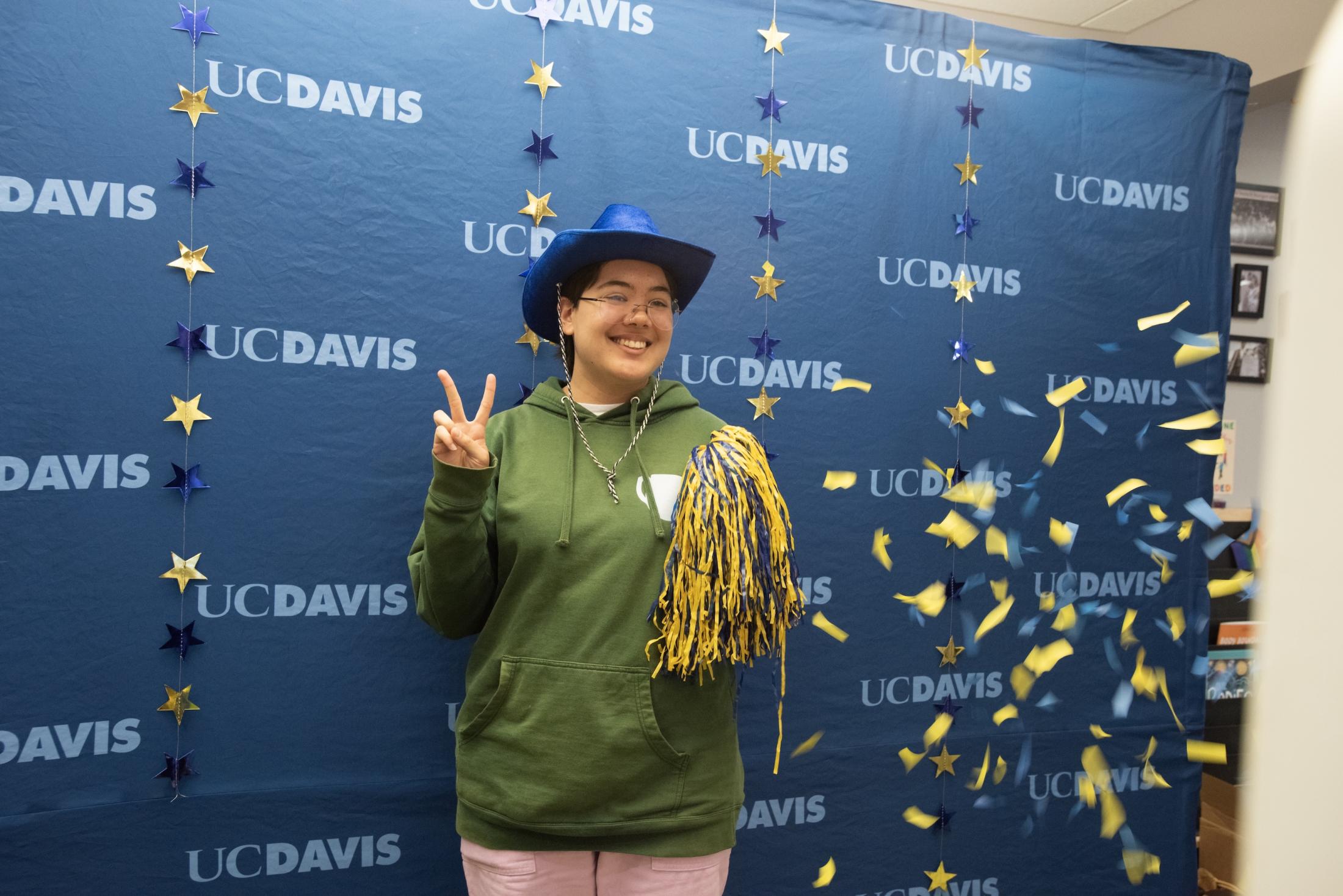 A UC Davis admit in a green hoodie and blue cowboy hat holding a blue and gold pompom, poses in front of a blue UC Davis backdrop decorated with stars. Confetti fills the air around them.