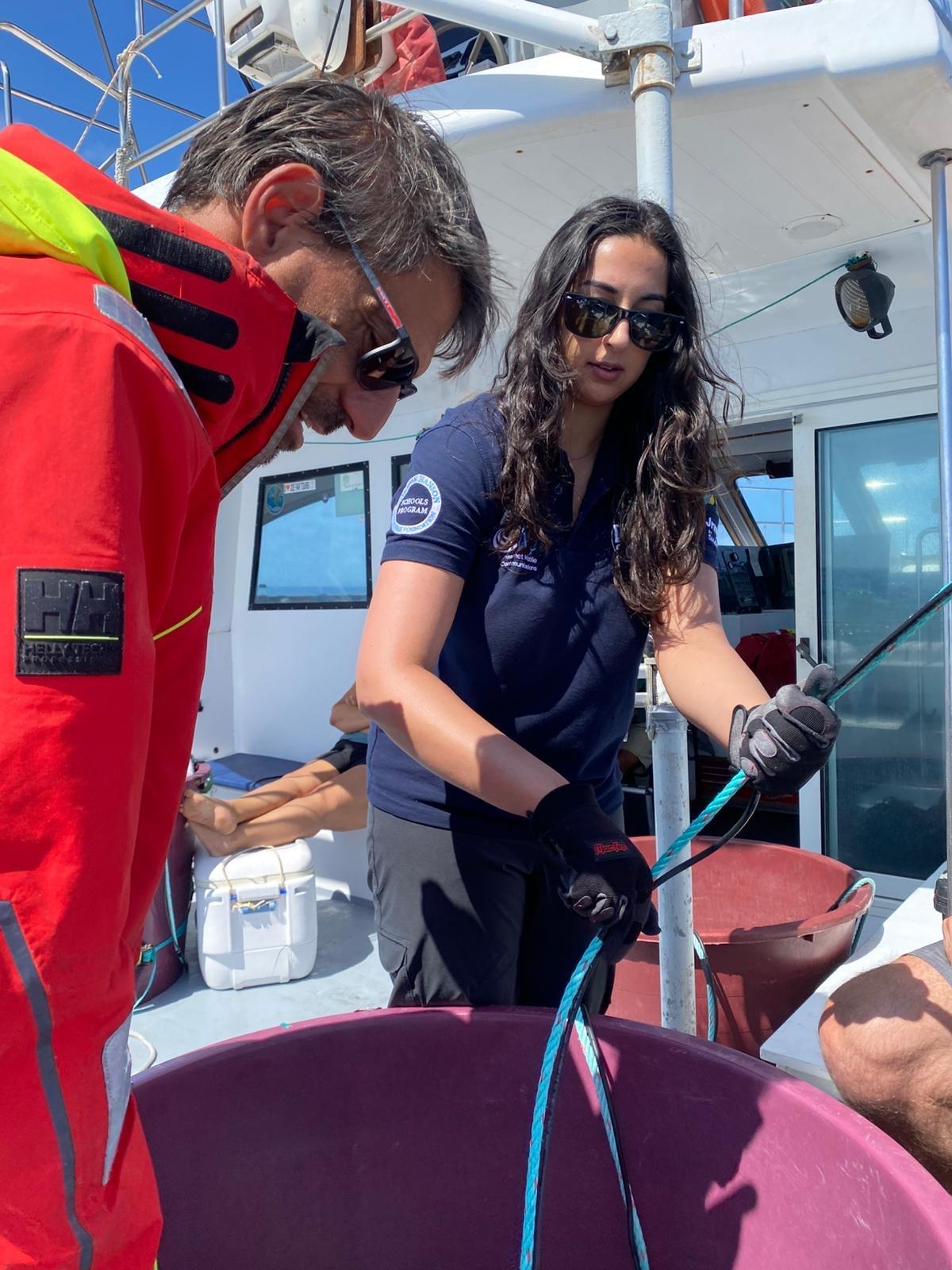Priya Talreja, wears sunglasses and gloves and works with rope aboard a research vessel alongside another crew member in a red jacket.