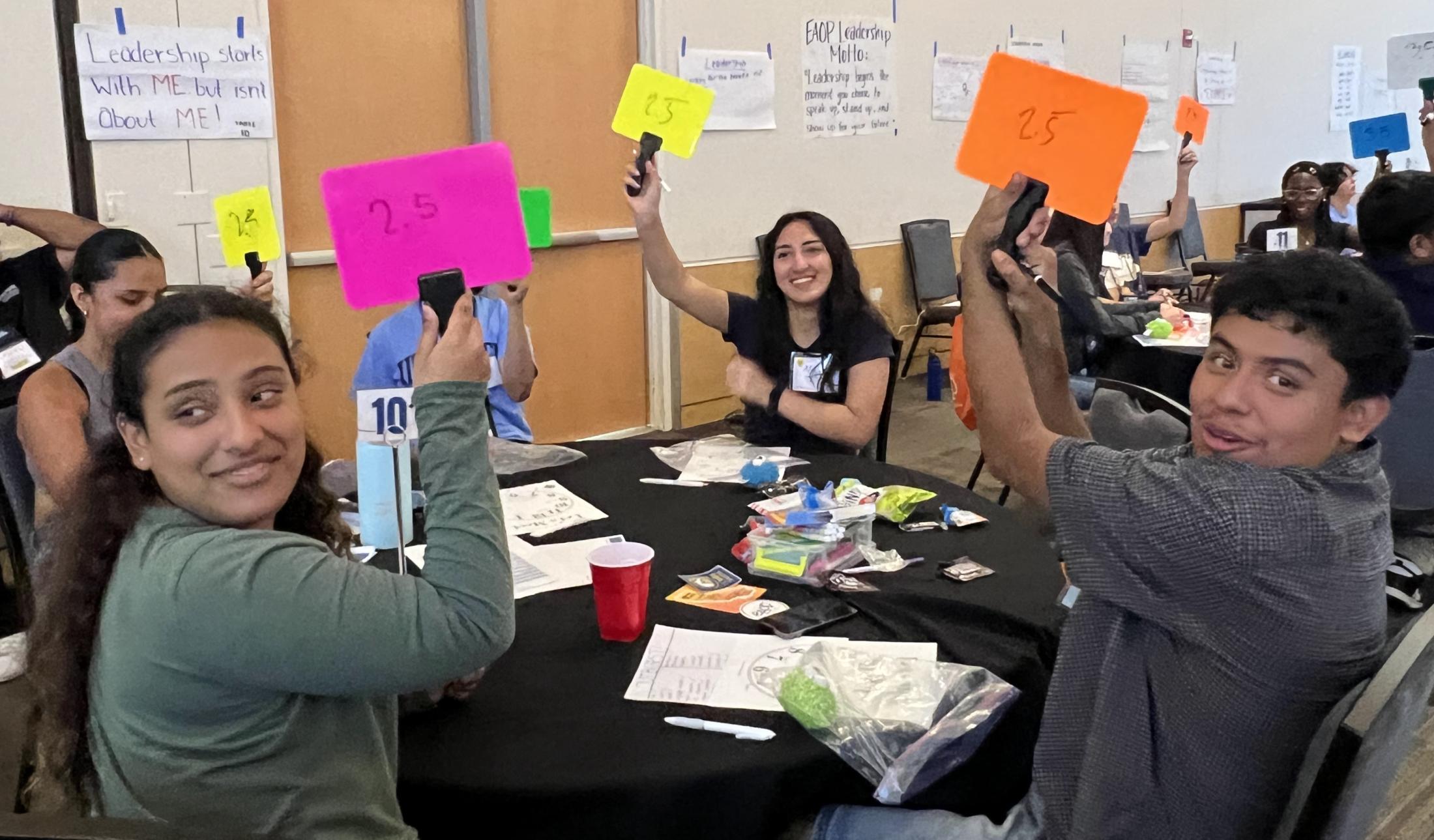 High school students sit around tables and hold up neon-colored paddles during a quiz game inside the UC Davis ARC Ballroom.