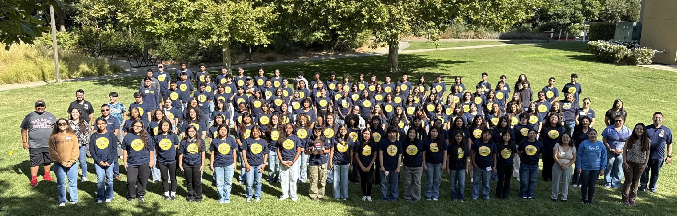 A large group of high school students and staff wear matching navy shirts and stand together on a lawn surrounded by trees.