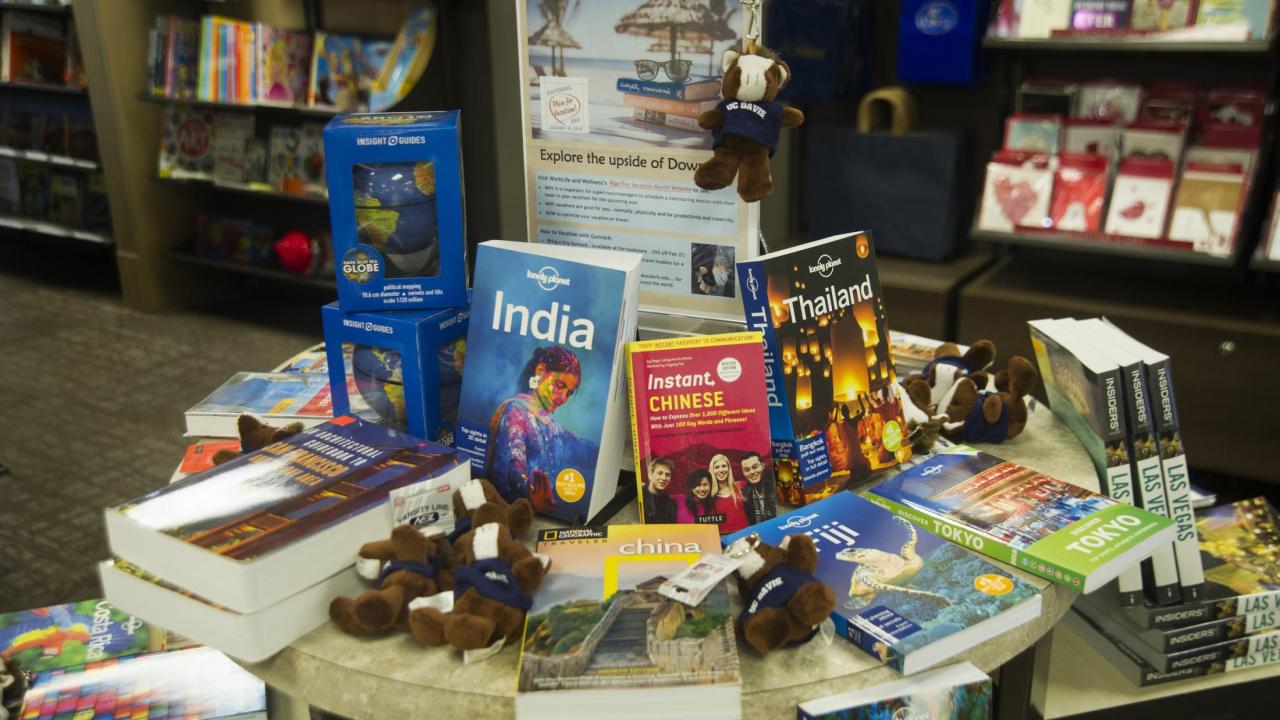Table display at the UC Davis bookstore with travel books on countries like India, Thailand and China. Several UC Davis Gunrock plush toys are scattered among the books.