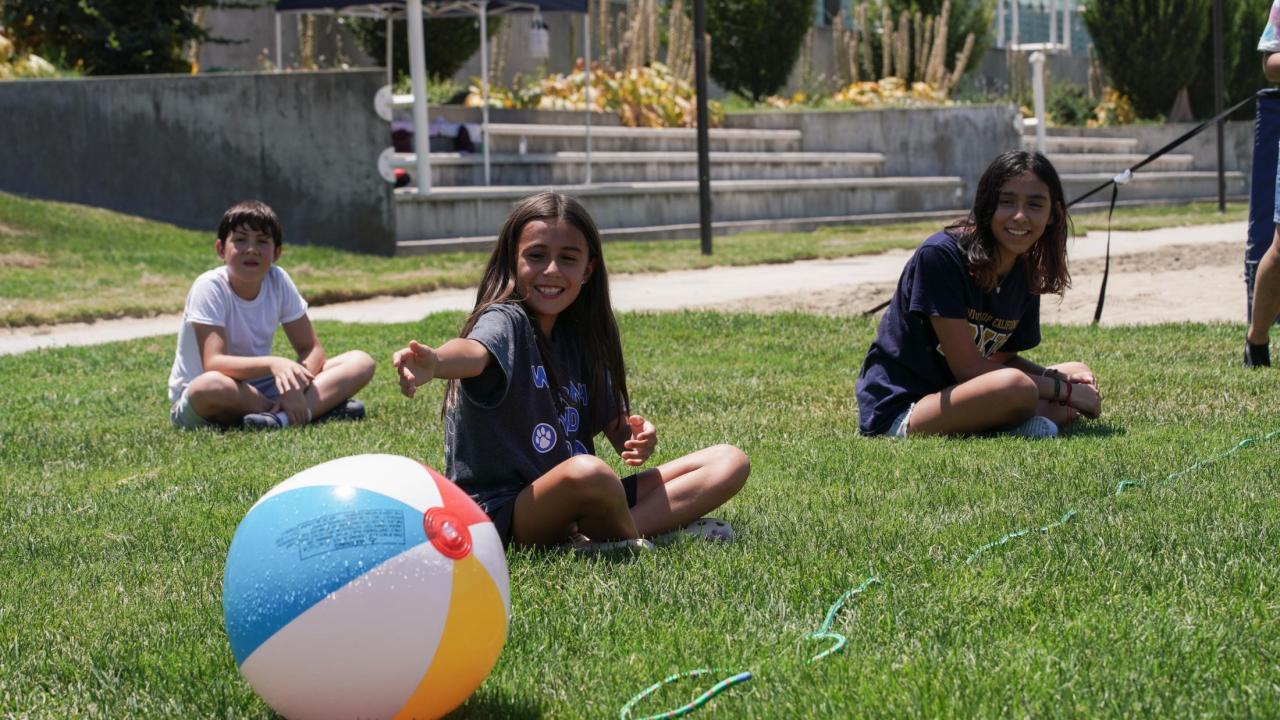 Three students sitting on grass looking over at a colorful beach ball.