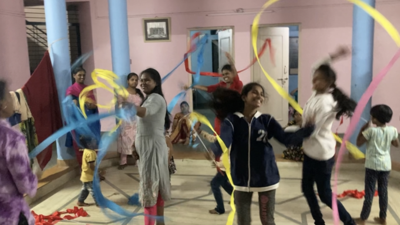 A group of children and women dance indoors, waving long colorful ribbons in the air. The room has light blue pillars, pink walls and a tiled floor.