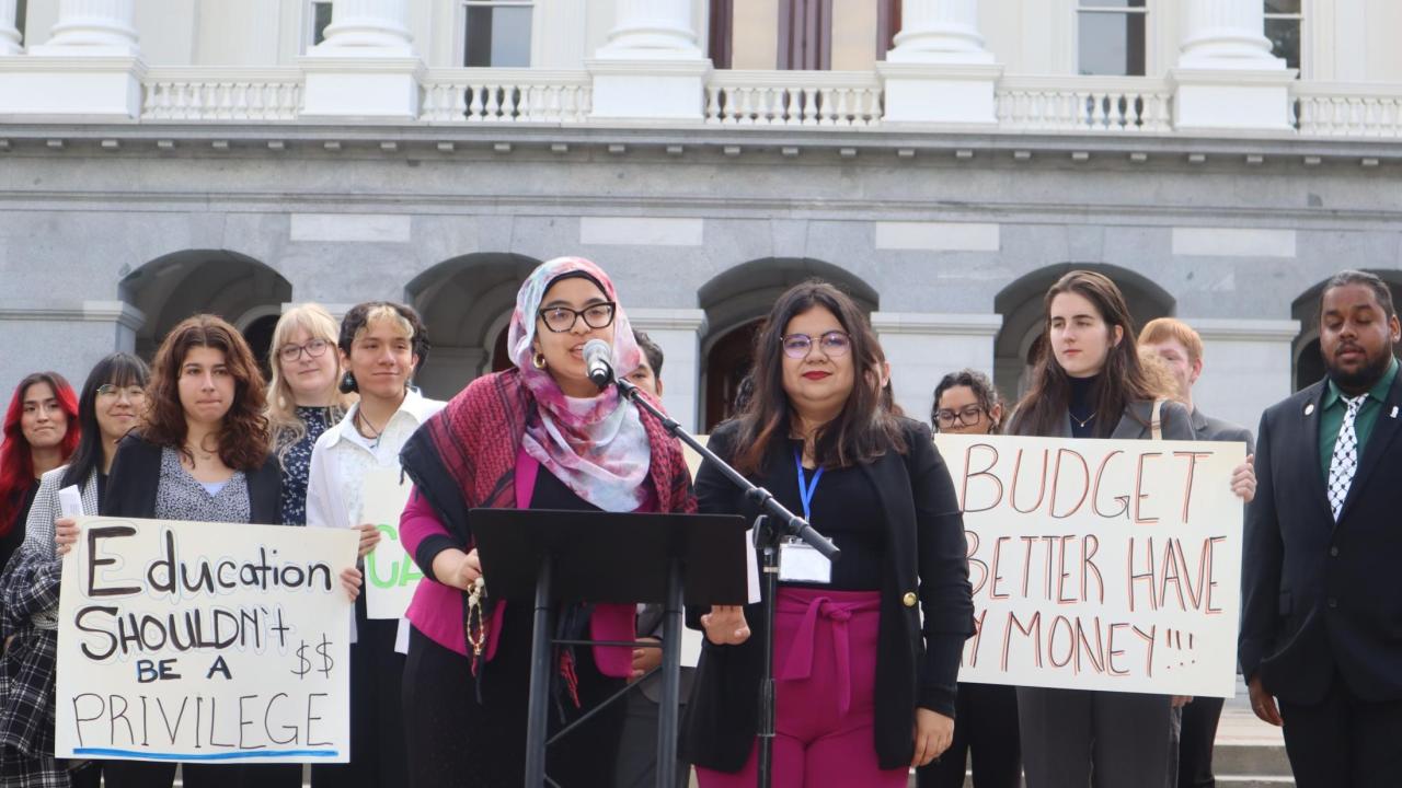 Aridin speaks at a podium in a hot pink outfit in front of a government building, surrounded by colleagues with signs that read “Education shouldn’t be a privilege” and “Budget better have my money.”