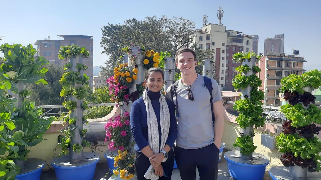 Two people stand on a rooftop garden surrounded by vertical hydroponic towers filled with leafy greens and flowers, with city buildings in the background on a sunny day.