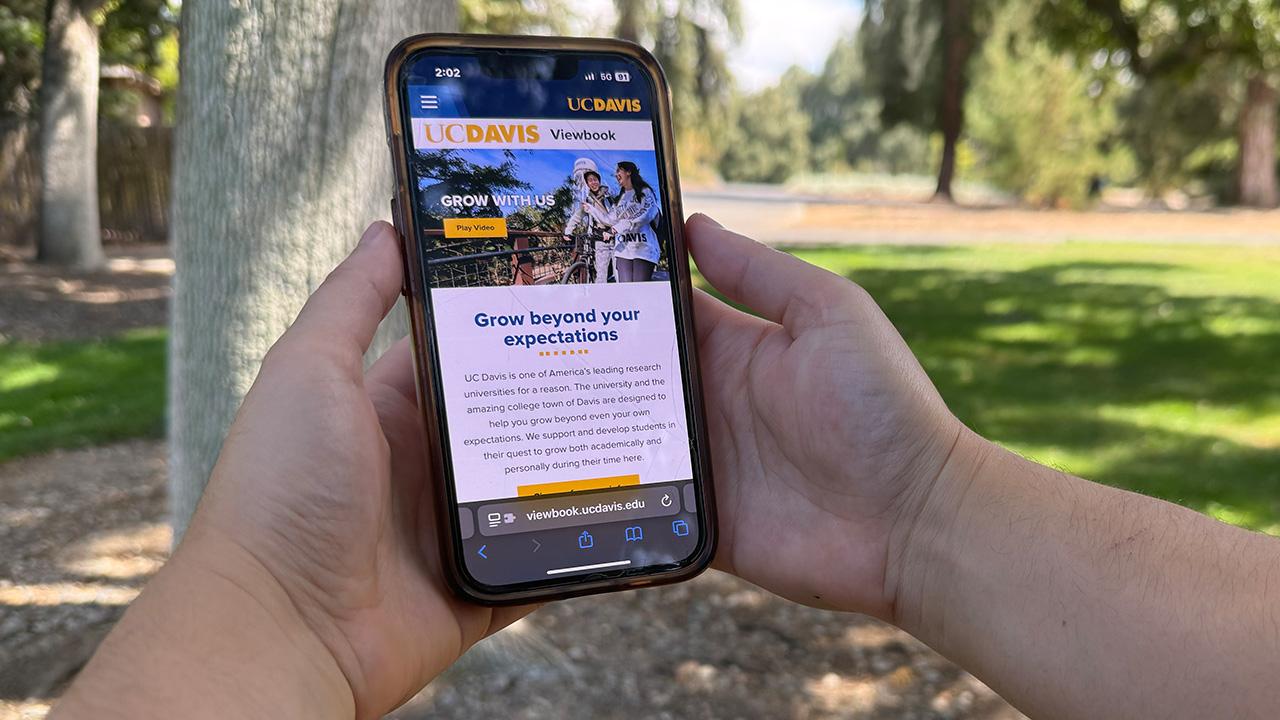 Two hands hold a mobile device displaying the website for the UC Davis viewbook against a blurred background of a grassy area.