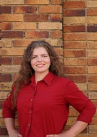 Emily Granger stands in front of a brick wall and smiles with her hands on her hips. She wears a red button-down shirt.