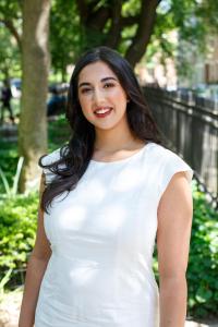 Priya Talreja stands outside in a shaded area with trees and greenery. She wears a white cap-sleeve dress and smiles at the camera.