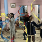 A group of children and women dance indoors, waving long colorful ribbons in the air. The room has light blue pillars, pink walls and a tiled floor.