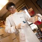 Undergraduate researcher wears safety goggles and gloves pours red liquid into a spray bottle labeled “70% EtOH Flammable” inside a laboratory workspace with shelves and equipment in the background.