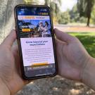 Two hands hold a mobile device displaying the website for the UC Davis viewbook against a blurred background of a grassy area.